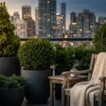 A cozy, green balcony sanctuary with various evergreen pots and a wooden chair overlooking a city at sunset.