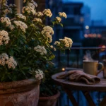 A glowing white flowering shrub in a terracotta pot on a small city balcony at dusk next to a wooden table and a warm candle.